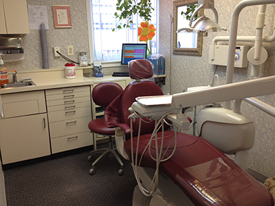 The image shows an interior view of a dental office with a chair in the foreground, dental equipment and tools, a desk with a computer monitor, and various other dental supplies visible in the background.