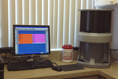 The image shows a desktop computer setup on a table with a monitor, keyboard, and mouse. In front of the computer, there s a large piece of equipment that appears to be a scientific or medical device, possibly for measuring or analyzing samples. To the right, there is a printer and what looks like a coffee maker. The setting seems to be a laboratory or research environment, with a window in the background showing blinds.