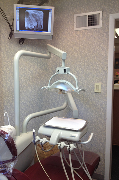 The image shows a dental office setting with a dental chair, a dental mirror, a monitor displaying an X-ray image, and a dental treatment table with various dental instruments on it.