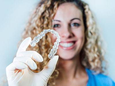 A woman holding up a transparent plastic dental retainer with both hands, smiling at the camera.