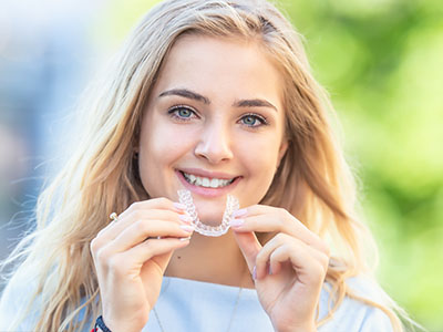 The image shows a woman smiling at the camera while holding a clear orthodontic appliance, possibly an aligner, between her teeth.