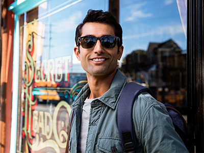 The image shows a man standing outdoors, wearing sunglasses and a backpack, with a cheerful expression, posing for a photo in front of a storefront with a sign that reads  Sweet Treats.