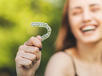 The image shows a woman holding a clear plastic toothbrush with bristles, smiling and looking towards the camera.