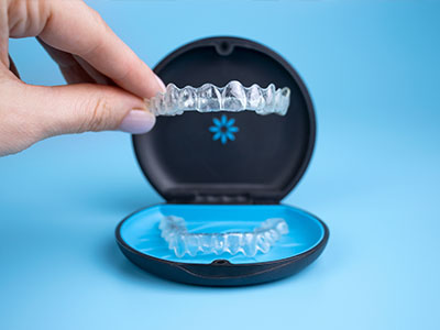 The image shows a hand holding an open box containing a clear tray with a set of transparent dental aligners, which are part of a teeth straightening system.