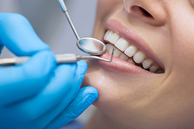 The image depicts a dental professional performing a teeth cleaning procedure on a patient s mouth using specialized equipment, with the patient wearing protective gloves and a blue surgical gown.