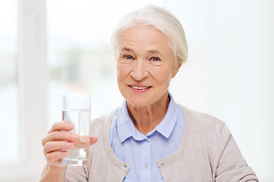 An elderly woman holding a glass of water, smiling at the camera with her hand raised slightly above the glass.