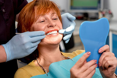 A woman sitting in a dental chair with a smile on her face, holding a blue mouthguard while a dentist adjusts it with tools.