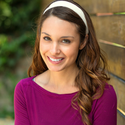 A young woman with long brown hair and a wide smile, wearing a purple top and a headband, posing against a wooden fence.