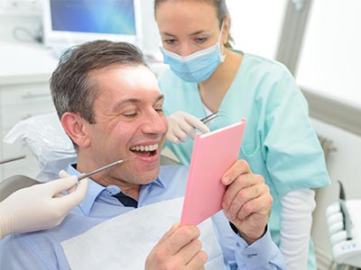 A man seated in a dental chair, smiling at a pink card in his hands, while a woman in scrubs looks on with a surgical mask covering her mouth.