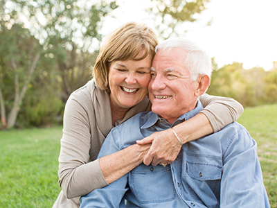 The image shows an elderly couple embracing each other outdoors during daylight, with both individuals smiling and appearing content.