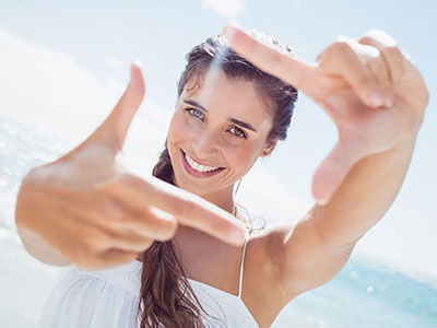 A woman with long hair, smiling at the camera, holds up a hand with fingers spread wide against a bright blue sky background.